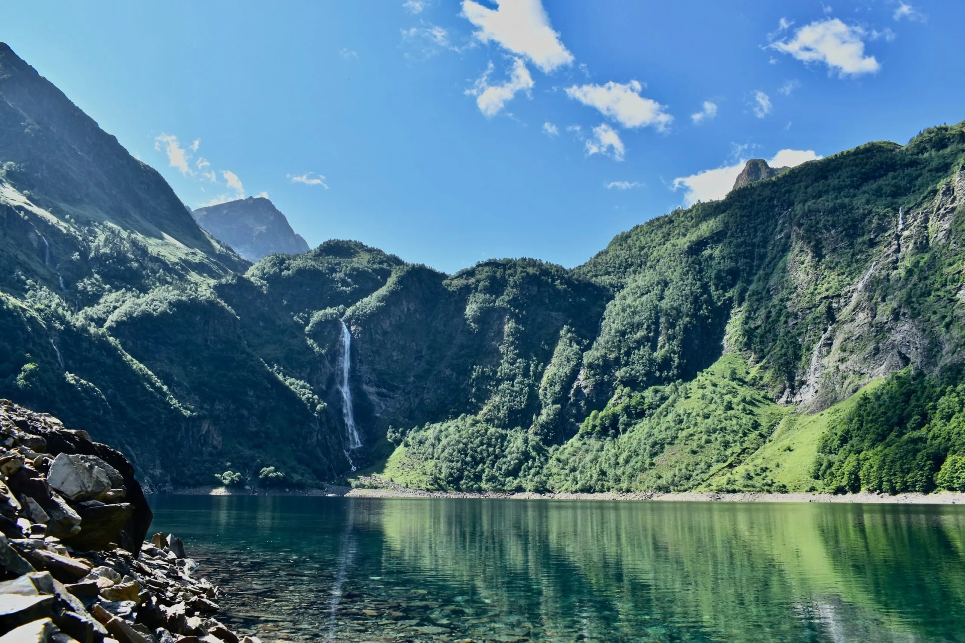 Waterfall cascading down lush green mountainsides into a clear alpine lake under a blue sky.